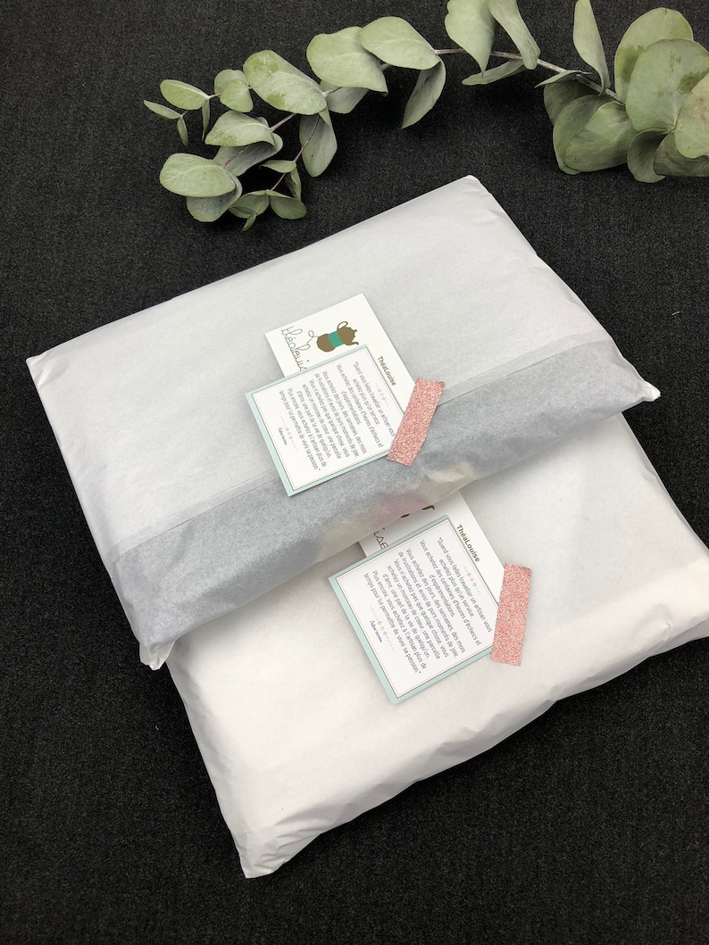 K&ouml;nnte beinhalten: Zwei wei&szlig;e Seidenpapierpakete mit einem rosa-wei&szlig;en Etikett mit der Aufschrift "These LAE" und einem blau-wei&szlig;en Etikett mit der Aufschrift "Thealouise".