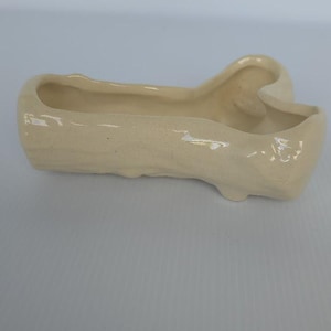 May include: A small, cream-colored ceramic planter with a unique, curved design. The planter has a long, rectangular trough with a raised, heart-shaped end. The surface has a textured appearance.