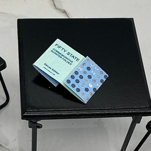 May include: A miniature scene featuring a black table with two small black chairs. On the table is a light blue and silver "Fifty State Commemorative Quarter Folder" booklet. The booklet is open, revealing a pattern of silver and blue circles.