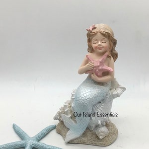 May include: A light blue mermaid figurine with a pink starfish in her hands, sitting on a bed of white seashells. The figurine is on a white background with a light blue starfish in the foreground.  Out Island Essentials.