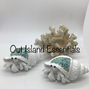 May include: Two white ceramic hermit crab figurines with blue and green mosaic shells. The figurines are sitting on a white surface.  Out Island Essentials is written in black text on the surface.