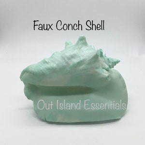 May include: A faux conch shell painted in a light blue colour. The shell is sitting on a white surface. The text "Faux Conch Shell" is above the shell and the text "Out Island Essentials" is below the shell.