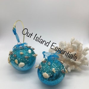 May include: Two blue glitter Christmas ornaments decorated with seashells and white beads. The ornaments are hanging from a gold wire hanger. The text "Out Island Essentials" is visible in the image.