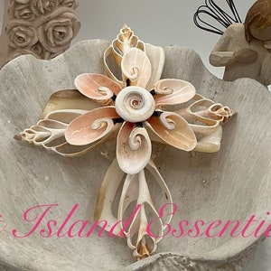 May include: A white seashell bowl with a seashell cross inside. The bowl is decorated with a white and gray marbled pattern. The cross is made of natural seashells and has a light pink and white color. The bowl is sitting on a white surface. "Out Island Essentials" is written on the bowl.