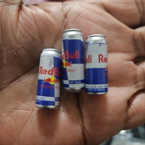 May include: Three miniature Red Bull energy drink cans. The cans are silver with blue and red branding, featuring the Red Bull logo and product information. The cans are small, likely for novelty or promotional purposes.