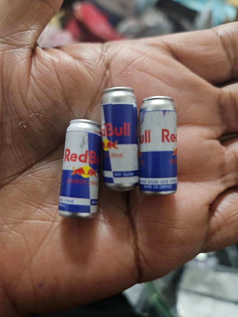 May include: Three miniature Red Bull energy drink cans. The cans are silver with blue and red branding, featuring the Red Bull logo and product information. The cans are small, likely for novelty or promotional purposes.