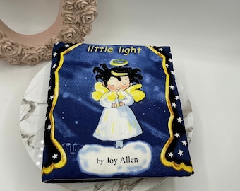 Pode incluir: Uma capa de livro azul com uma borda amarela e estrelas brancas. A capa apresenta uma ilustração de um anjo com cabelo escuro e uma auréola segurando uma vela. O título do livro é "little light" e o autor é "by Joy Allen".