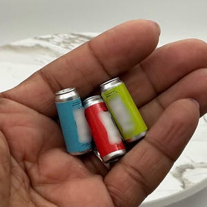 May include: Three miniature soda cans in blue, red, and yellow, held in a hand. The cans have silver tops and white labels. The background is a white marble-patterned surface.