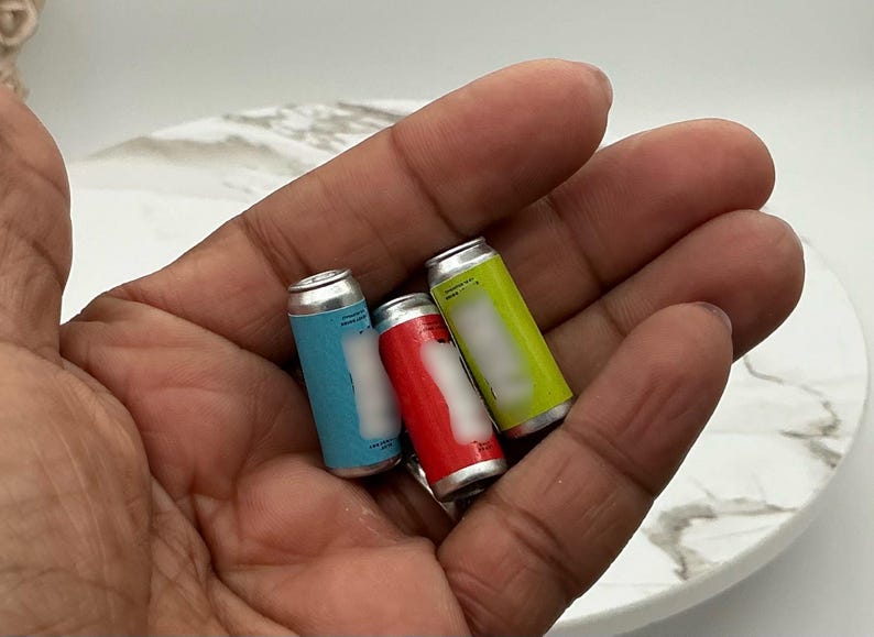 May include: Three miniature soda cans in blue, red, and yellow, held in a hand. The cans have silver tops and white labels. The background is a white marble-patterned surface.