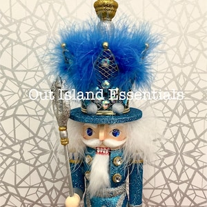 May include: A decorative nutcracker in a teal blue uniform with silver accents, a white beard, and a tall hat adorned with blue feathers and a gold crown. The text "Out Island Essentials" is visible.