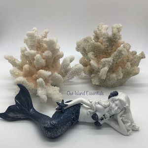 Pu&ograve; includere: Una statuetta di sirena bianca con una coda blu e una stella marina in mano, sdraiata su uno sfondo bianco con due pezzi di corallo bianco dietro di lei.  Out Island Essentials &egrave; scritto sullo sfondo.