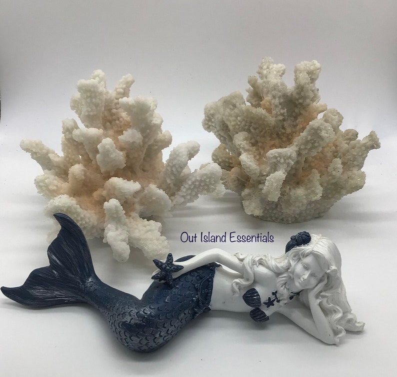 Pu&ograve; includere: Una statuetta di sirena bianca con una coda blu e una stella marina in mano, sdraiata su uno sfondo bianco con due pezzi di corallo bianco dietro di lei.  Out Island Essentials &egrave; scritto sullo sfondo.
