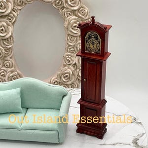 May include: A miniature wooden grandfather clock with a brown finish and a gold face. The clock is standing on a white surface with a light blue sofa in the background.