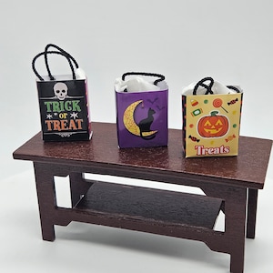 May include: Three miniature Halloween treat bags on a brown wooden table. The bags are decorated with different Halloween themes: a skull with the text "Trick or Treat", a black cat with a crescent moon, and a jack-o'-lantern with the text "Treats".