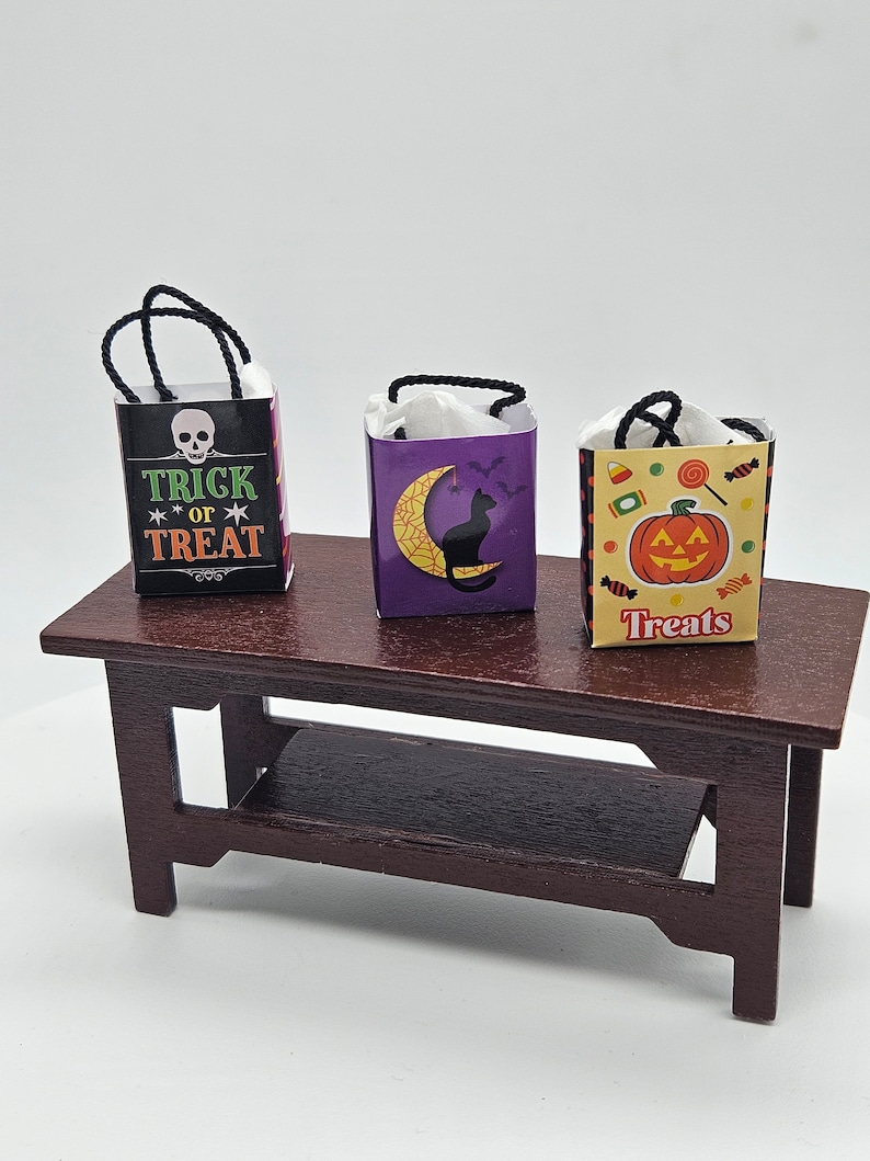 May include: Three miniature Halloween treat bags on a brown wooden table. The bags are decorated with different Halloween themes: a skull with the text "Trick or Treat", a black cat with a crescent moon, and a jack-o'-lantern with the text "Treats".