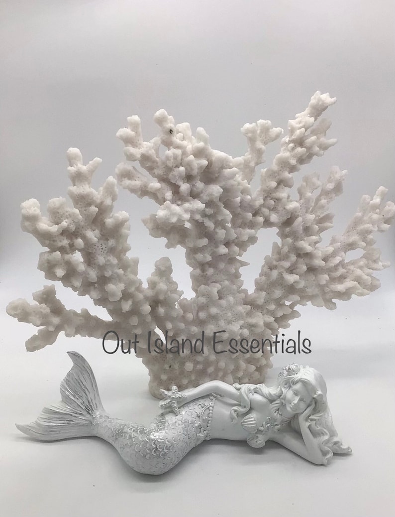 Pu&ograve; includere: Figurina di sirena in ceramica bianca con una lunga coda fluida, sdraiata su una barriera corallina bianca. La sirena indossa una corona di fiori sulla testa e tiene una stella marina in mano. Il testo "Out Island Essentials" &egrave; visibile nell'immagine.