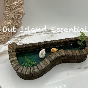 Puede incluir: Una escena de estanque en miniatura con un borde de ladrillo, con un gato, un cisne blanco y dos patitos amarillos. El estanque tiene agua verde y plantas acuáticas. El texto "Out Island Essentials" es visible.
