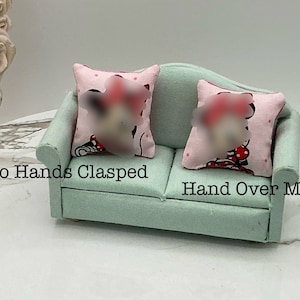 May include: A light green miniature sofa with two pink and white throw pillows featuring a cartoon character. The pillows have the text "Two Hands Clasped" and "Hand Over Mouth".