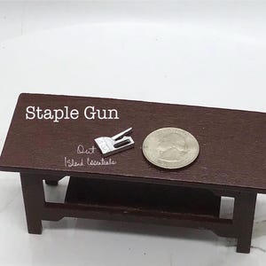 May include: A silver miniature staple gun on a dark brown wooden table with a silver quarter on the table. The text "Out Island Essentials" is written on the table.