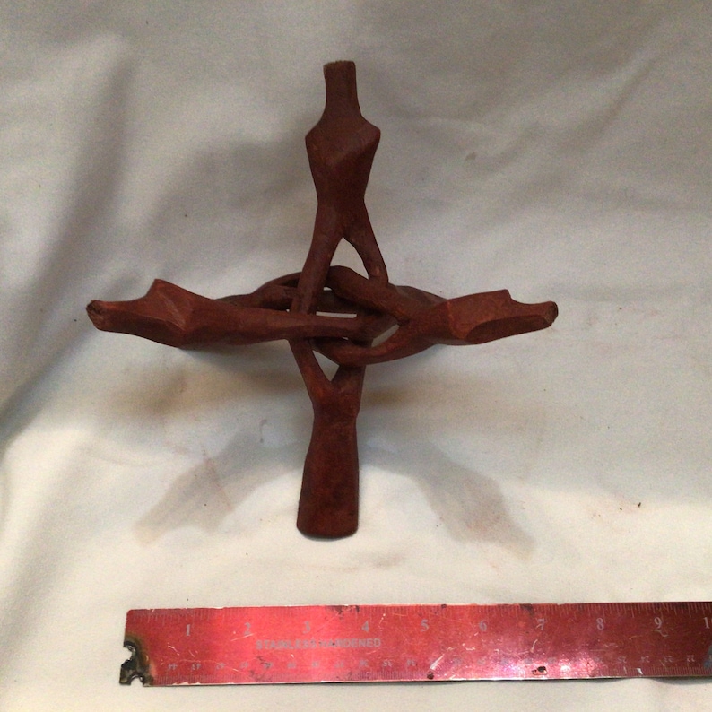 Op de afbeelding: Een houten sculptuur van vier in elkaar verweven handen, die een kruisvorm vormen. Het beeld is roodbruin van kleur en lijkt met de hand te zijn gesneden.