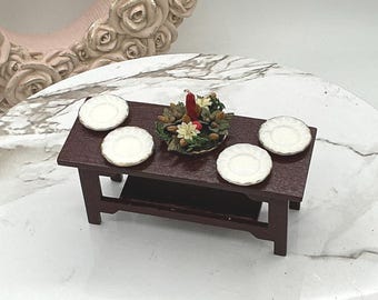May include: A miniature wooden table set with four white plates and a centerpiece of red candles and greenery.