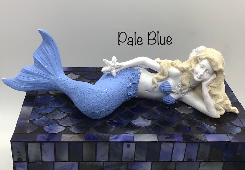 Pu&ograve; includere: Figurina di sirena blu pallido con un reggiseno di conchiglia bianca e una stella marina in mano. La sirena &egrave; sdraiata su una scatola di mosaico blu.