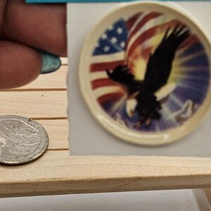 Op de afbeelding: Miniatuur ronde decoratieve plaat met een Amerikaans vlagontwerp met een zwarte adelaar in vlucht. De plaat staat op een kleine houten tafel, naast een munt voor de maatvergelijking.