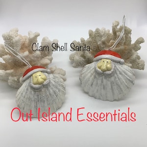 Puede incluir: Dos adornos de concha de almeja blanca decorados para parecerse a Papá Noel. Cada adorno tiene un gorro de Papá Noel rojo, barba blanca y cara amarilla. Los adornos cuelgan de coral blanco. El texto "Clam Shell Santa" y "Out Island Essentials" es visible en la imagen.