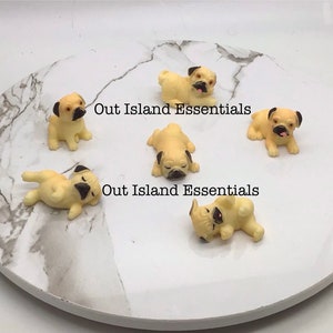 Puede incluir: Seis pequeñas figuritas de pug beige con ojos y narices negros. Las figuritas están dispuestas sobre una superficie de mármol blanco. El texto "Out Island Essentials" aparece dos veces en la superficie.