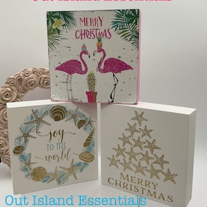 May include: Three wooden blocks with Christmas-themed designs. The first block features pink flamingos with the text "Merry Christmas". The second block has a blue and white wreath with seashells and the text "joy to the world". The third block has a gold star tree with the text "Merry Christmas".