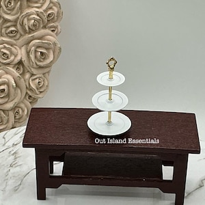 May include: A miniature three-tiered serving stand with white plates and a gold-coloured handle. It sits on a dark brown wooden table. The text "Out Island Essentials" is printed on the table.