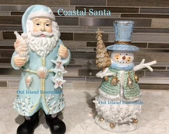 Pode incluir: Duas figuras decorativas: um Papai Noel costeiro e um boneco de neve costeiro. A figura do Papai Noel está em um casaco azul claro com detalhes de conchas, segurando conchas e estrelas do mar. O boneco de neve usa uma cartola e segura uma pequena árvore de Natal. O texto "Coastal Santa" e "Coastal Snowman" é visível.