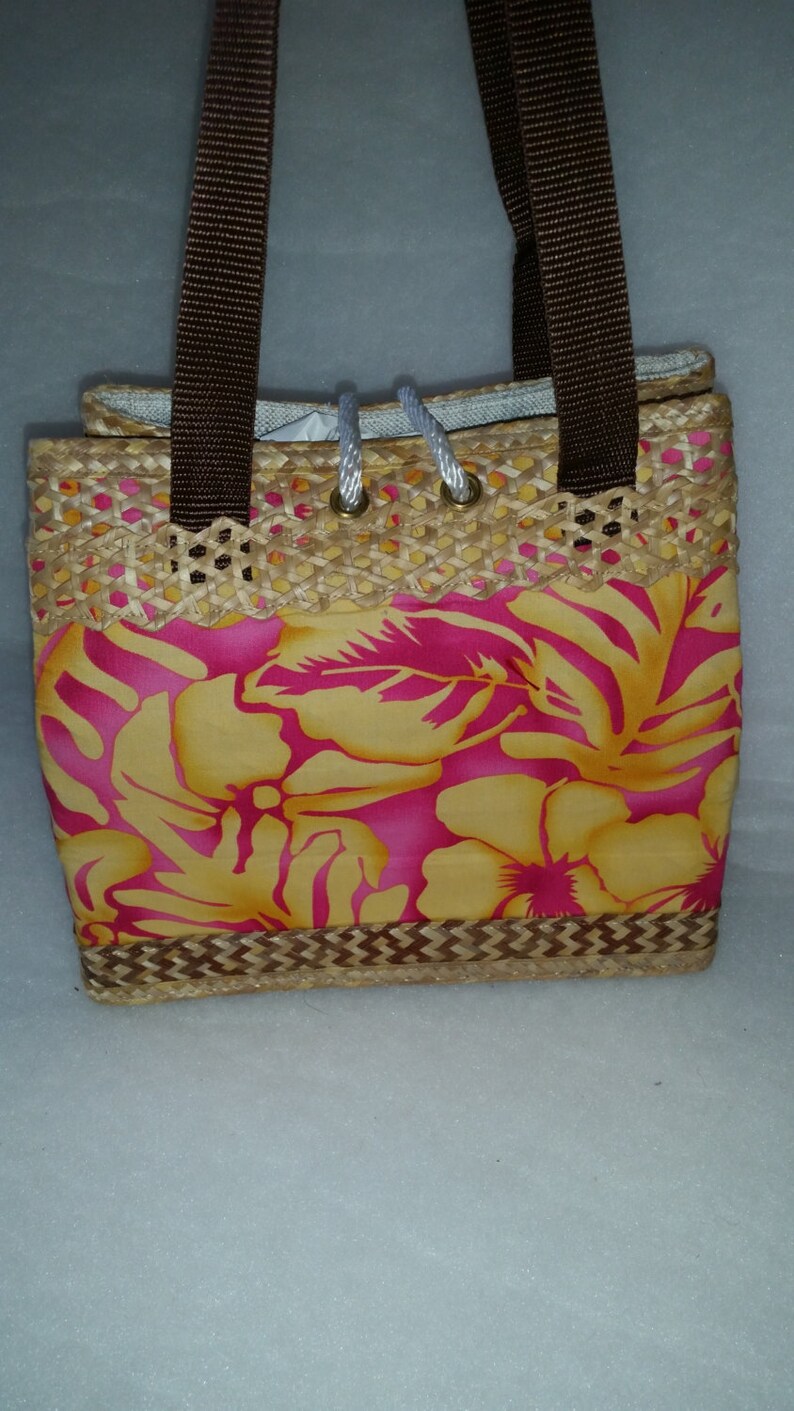 Puede incluir: Un peque&ntilde;o bolso de paja con asas marrones y estampado floral. El estampado presenta flores rosas y amarillas sobre un fondo amarillo.