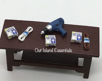 May include: A miniature table with a blue and silver cordless drill, two boxes of soft white light bulbs, and two spools of electrical wire, one white and one brown. The table has a brown wooden top and legs. The text "Out Island Essentials" is printed on the table.