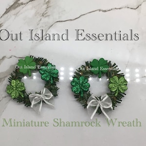 Puede incluir: Dos coronas de trébol en miniatura con tréboles de purpurina verde y lazos blancos. Las coronas están hechas de material verde y marrón. El texto "Out Island Essentials" y "Miniature Shamrock Wreath" es visible en la imagen.