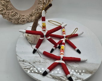 May include: Three red starfish ornaments painted to look like Santa Claus. Each starfish has a white beard, black eyes, and a yellow face. The starfish are on a white marble surface.