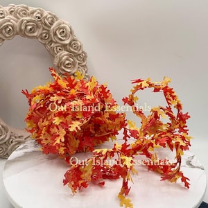May include: A display of artificial fall leaves in shades of red, orange, and yellow. The leaves are arranged in a bundle and a garland, with the text "Out Island Essentials" visible. A decorative floral wreath is in the background.