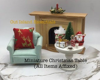 May include: A miniature Christmas table setting with a white table, a small Christmas tree, a Santa figurine, a gift box, a candy cane, and a glass of milk. The table is set on a marble surface in front of a miniature fireplace.