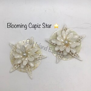 May include: Two white capiz shell star ornaments with a pearl in the center of each flower. The stars are on a gold glitter background.
