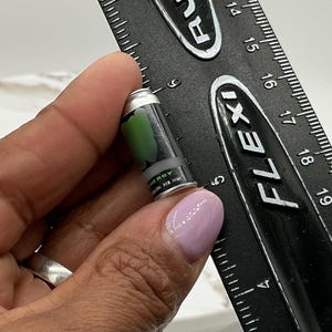 May include: A miniature black and silver energy drink can with a green design is held next to a black ruler. The can has the word "ENERGY" printed on it. The ruler is marked in inches.
