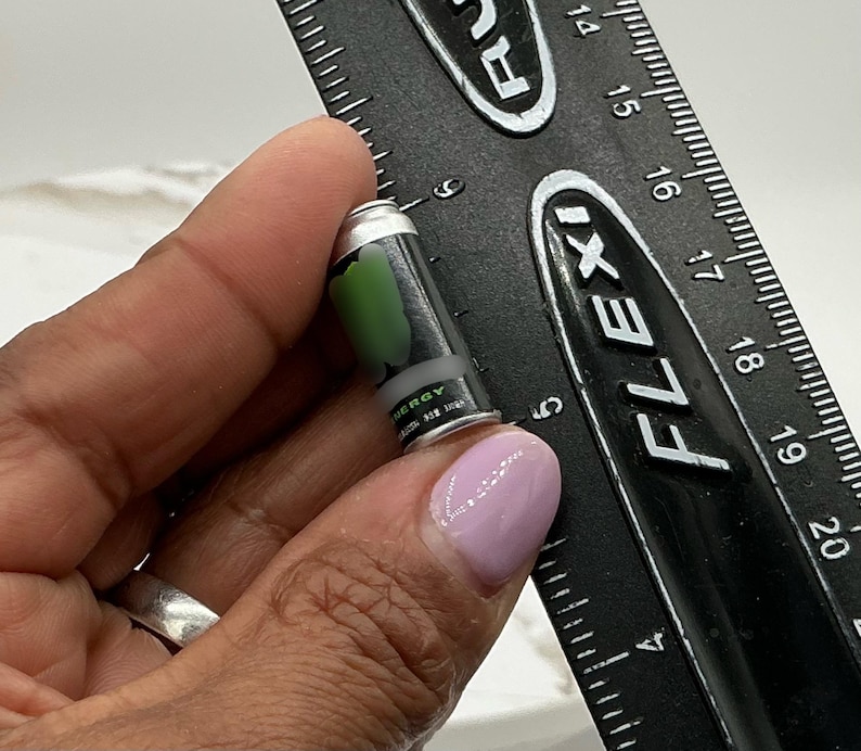 May include: A miniature black and silver energy drink can with a green design is held next to a black ruler. The can has the word "ENERGY" printed on it. The ruler is marked in inches.