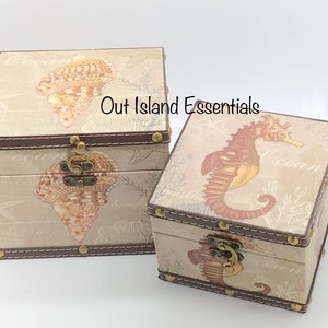 May include: Two decorative wooden boxes with a nautical theme. The larger box features a seashell design and the smaller box features a seahorse design. Both boxes have a brown leather trim and brass accents.