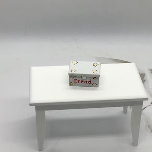 Puede incluir: Caja de pan de madera pintada de blanco con una etiqueta roja "Pan" y diseños de flores amarillas y rojas.