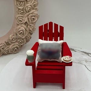 Puede incluir: Una silla Adirondack de madera roja con un cojín blanco, un vaso de papel con tapa y una dona en un plato blanco.