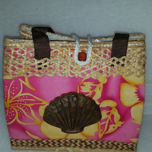 Puede incluir: Un bolso de mano floral rosa y amarillo con un dise&ntilde;o de concha marr&oacute;n. El bolso est&aacute; hecho de paja tejida y tiene asas marrones.