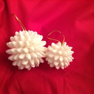 Puede incluir: Dos adornos de conchas blancas con colgadores de cuerda dorada. Los adornos tienen forma de bola y están hechos de muchas pequeñas conchas blancas.