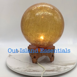 May include: A gold crackle glass globe lamp with a wooden base. The lamp is on a white marble surface. The text "Out Island Essentials" is visible in the image.