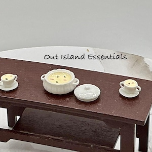 May include: A miniature dark brown wooden table featuring a white ceramic tureen with a matching lid, two small white ceramic cups on saucers, and the text Out Island Essentials printed in black above the items on the table surface.