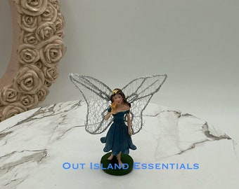 May include: A blue fairy figurine with silver wings holding a yellow flower. The fairy is standing on a green base.  Out Island Essentials is printed on the base.