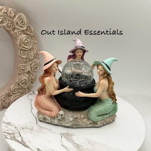 May include: A decorative figurine featuring three mermaids with long hair and pointed hats surrounding a central black cauldron with a cracked glass sphere. The text Out Island Essentials is visible above the figures, which sit on a base.